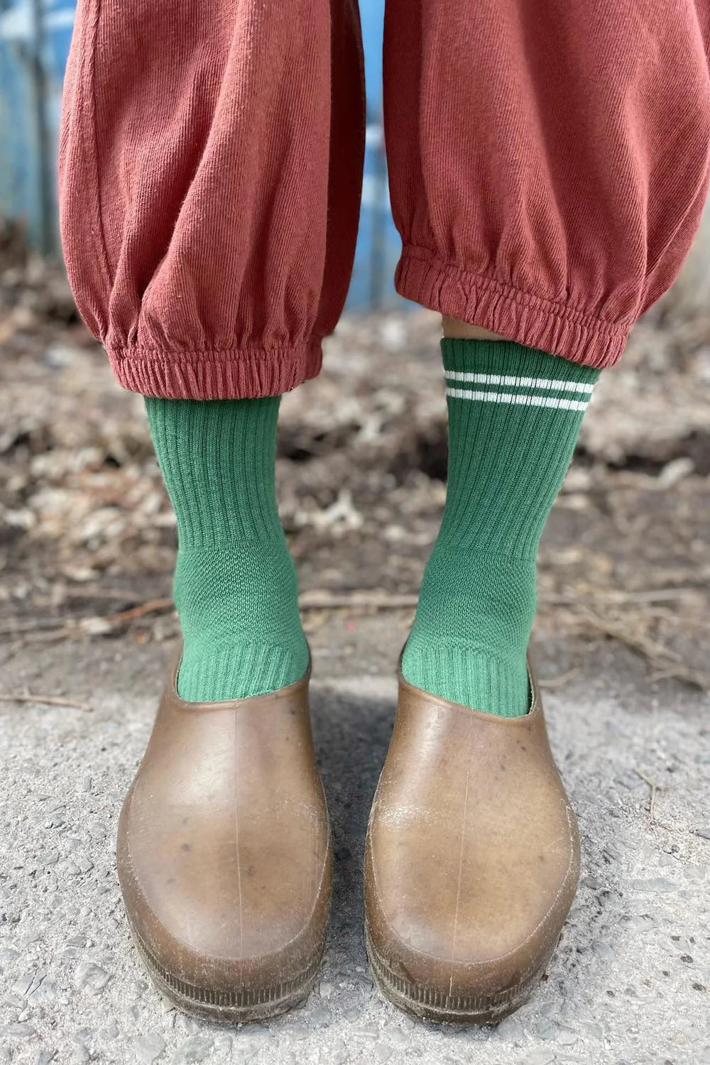Person wearing garden shoes, green socks with white stripes and pink pants standing on a sidewalk.