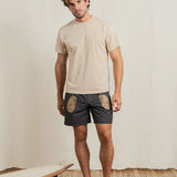 Man wearing a beige t-shirt and dark shorts with patterns, standing on a wooden floor with a white wall background.