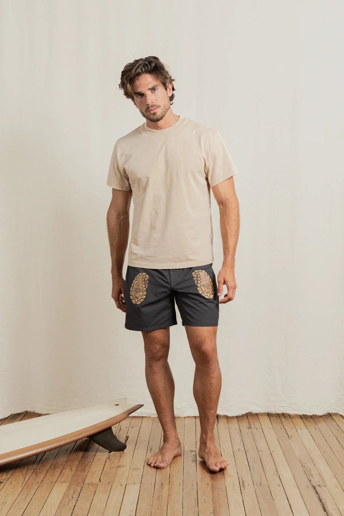 Man wearing a beige t-shirt and dark shorts with patterns, standing on a wooden floor with a white wall background.