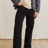 Person wearing black pants and a gray jacket on a wooden floor with a white wall background