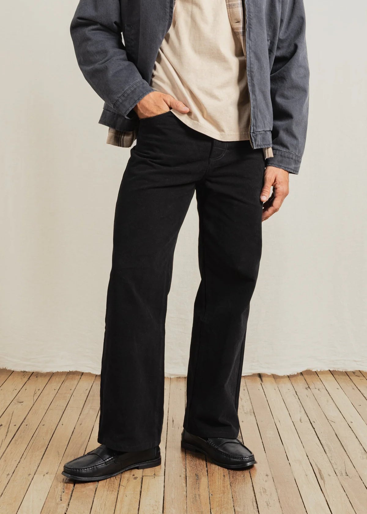 Person wearing black pants and a gray jacket on a wooden floor with a white wall background