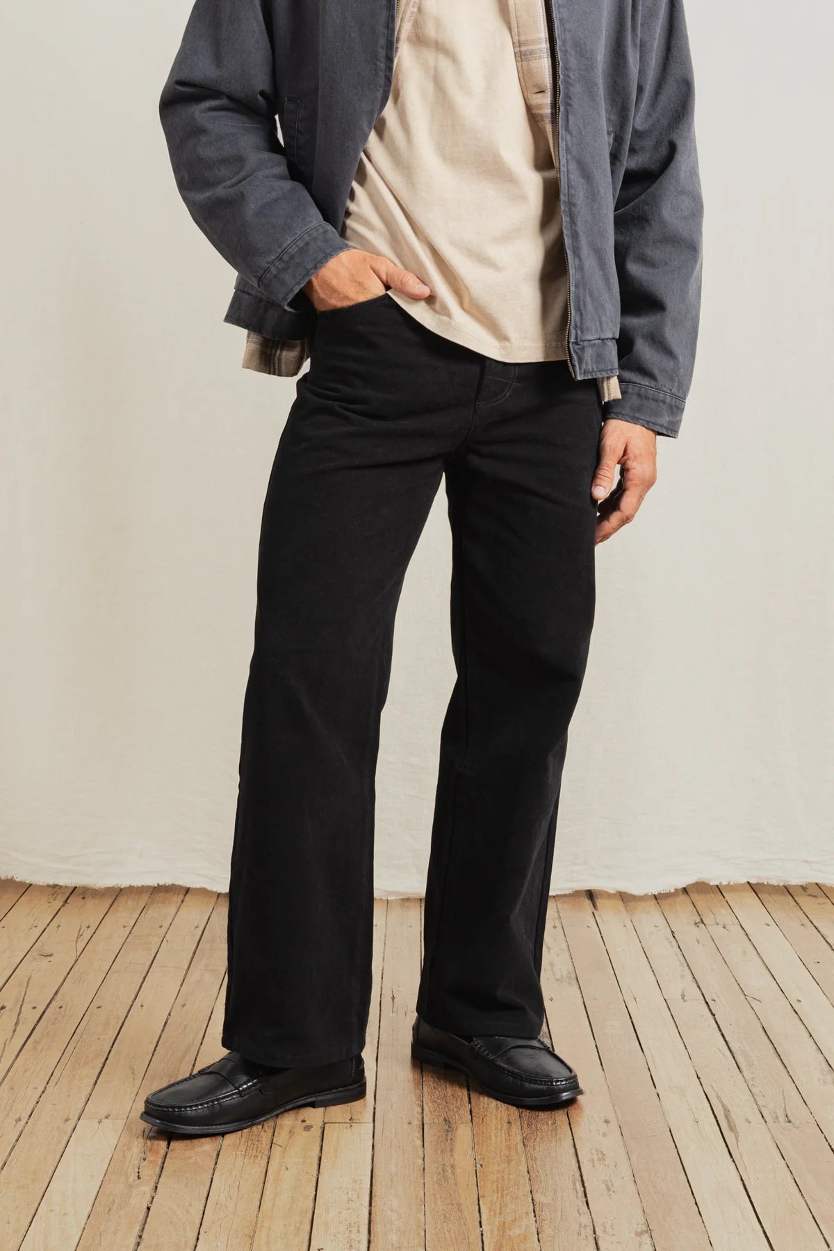 Person wearing black pants and a gray jacket on a wooden floor with a white wall background