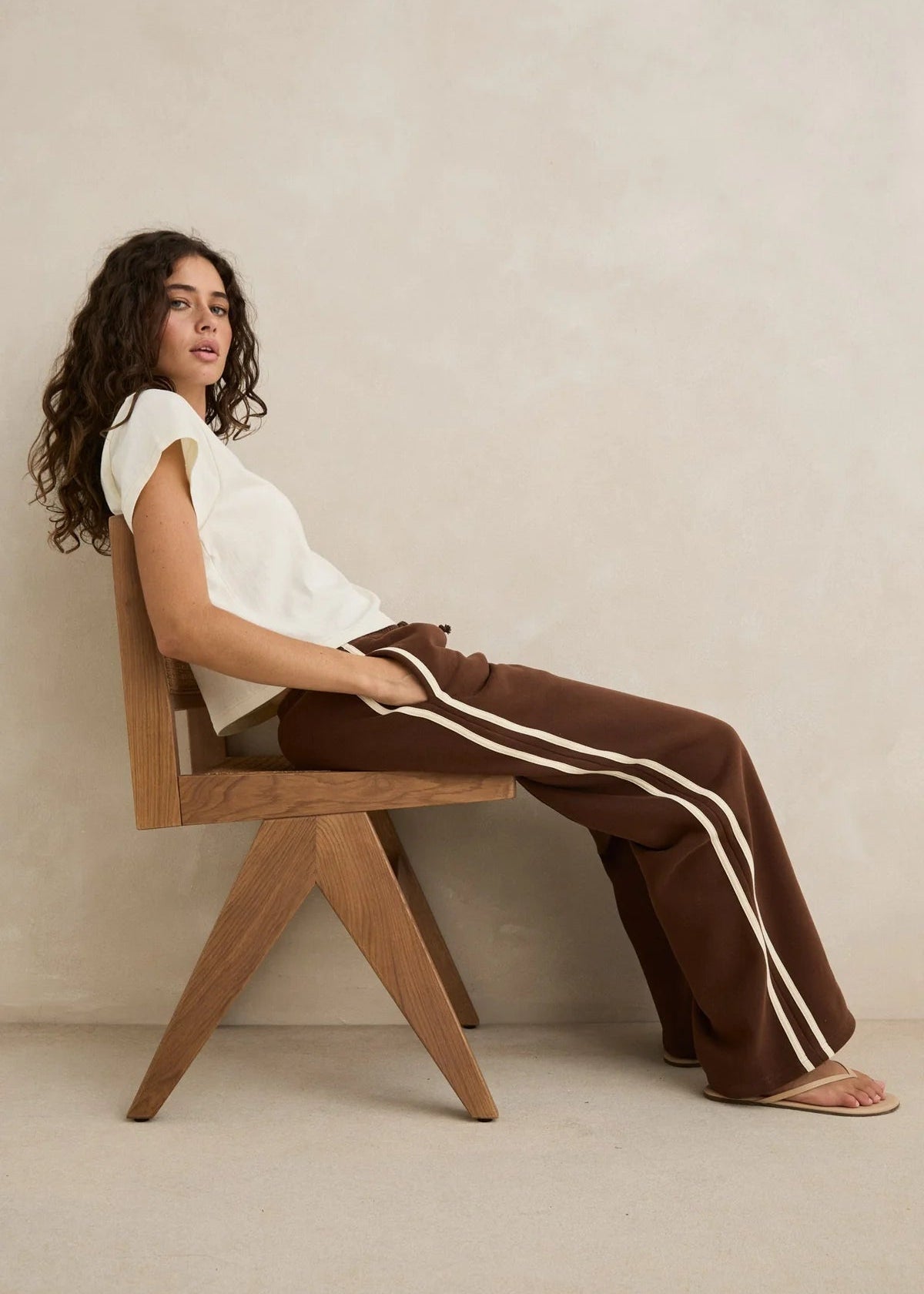 Woman sitting on a wooden chair wearing a white top and brown pants with white stripes.