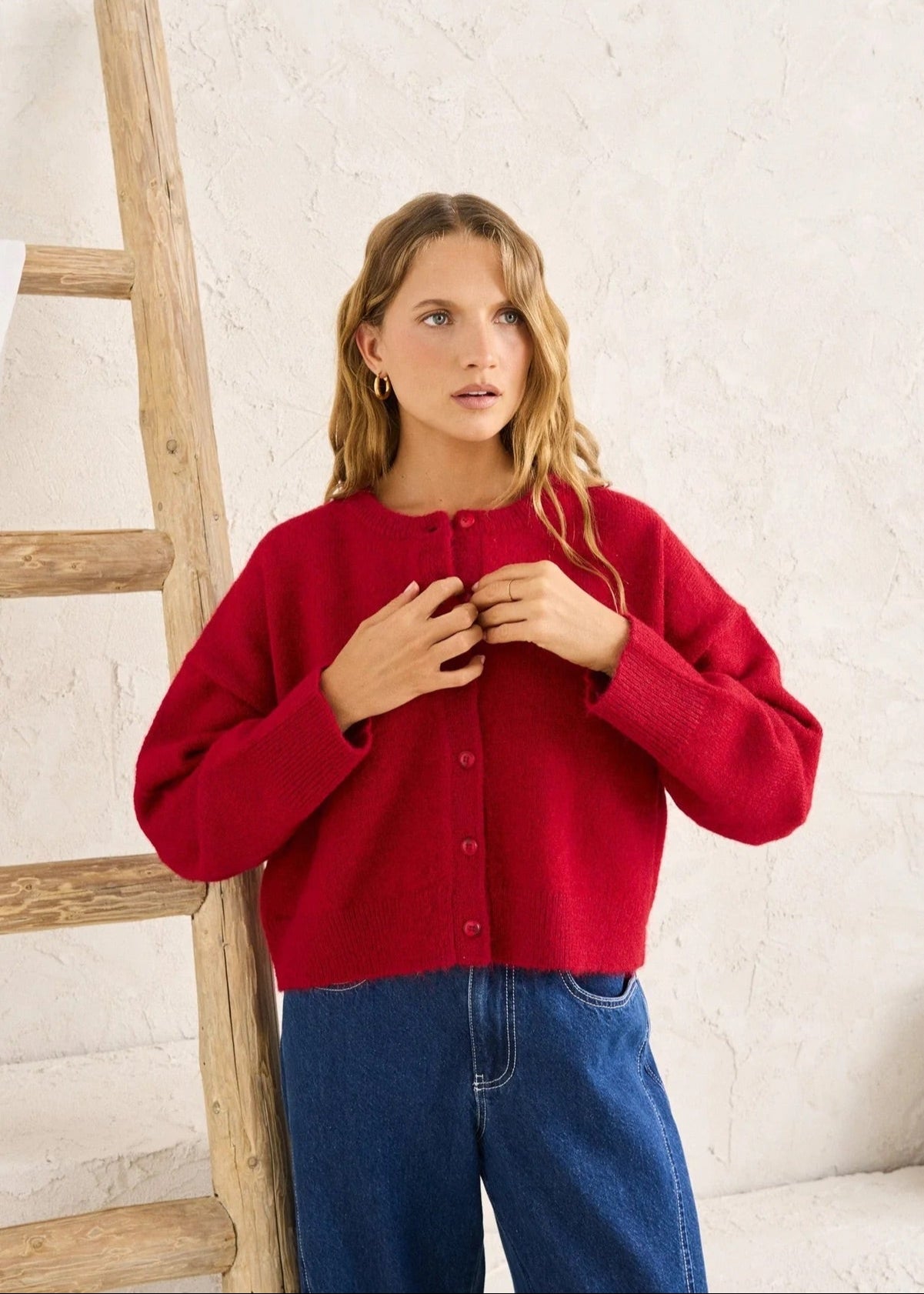 Woman wearing a red sweater and blue jeans standing next to a wooden ladder indoors.