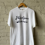 White t-shirt with 'Michigan Summers' text on a wooden hanger against a brick wall.