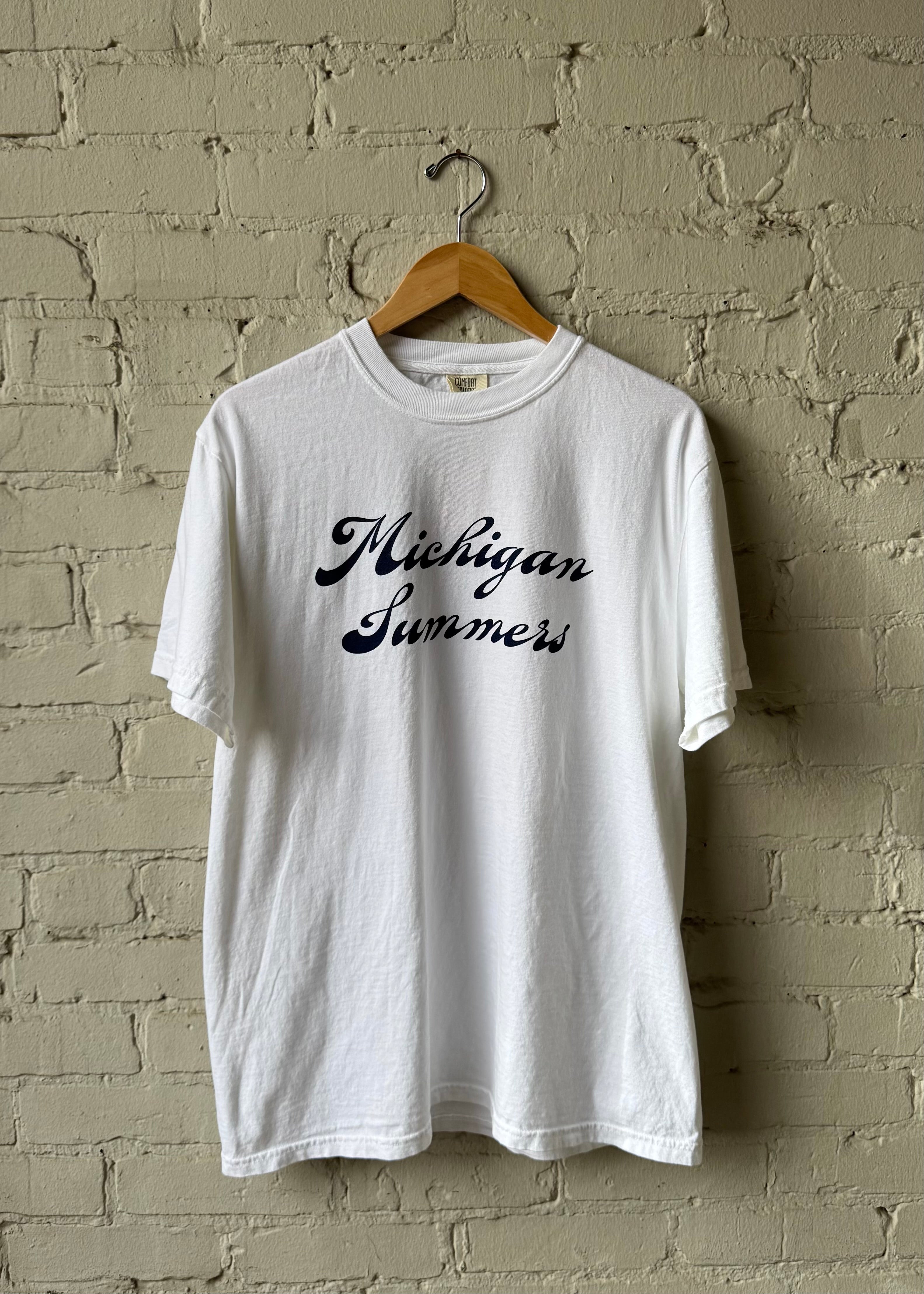 White t-shirt with 'Michigan Summers' text on a wooden hanger against a brick wall.