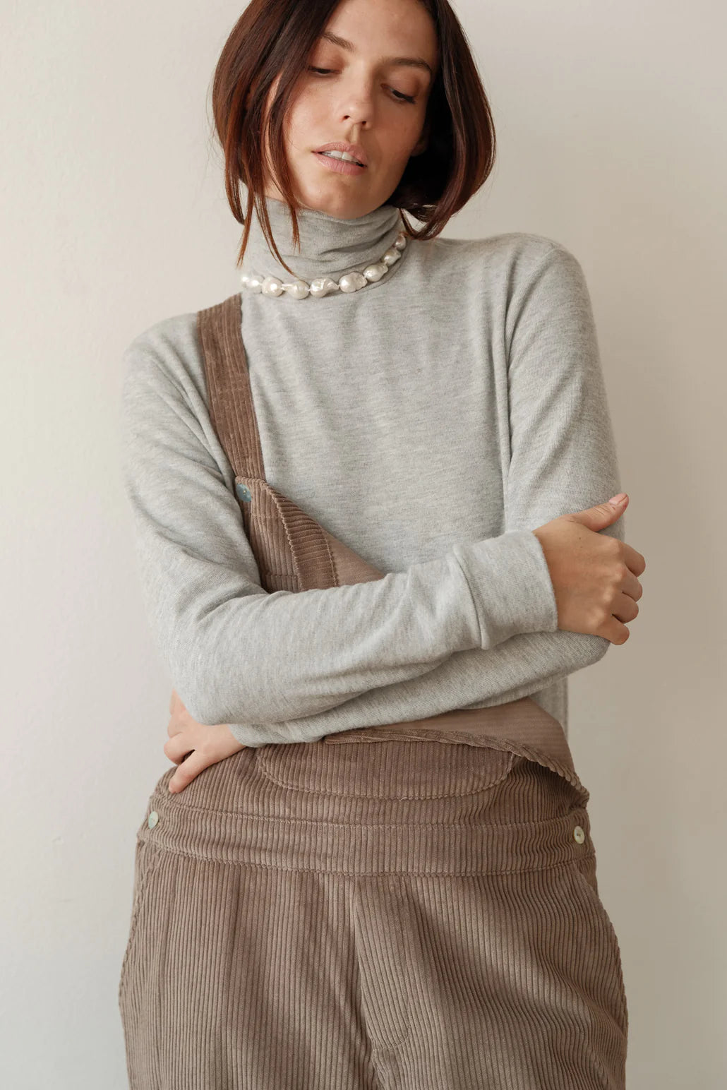 A close up image of a model posing and wearing the sweater turtleneck in heather grey styled under corduroy overalls and a pearl necklace