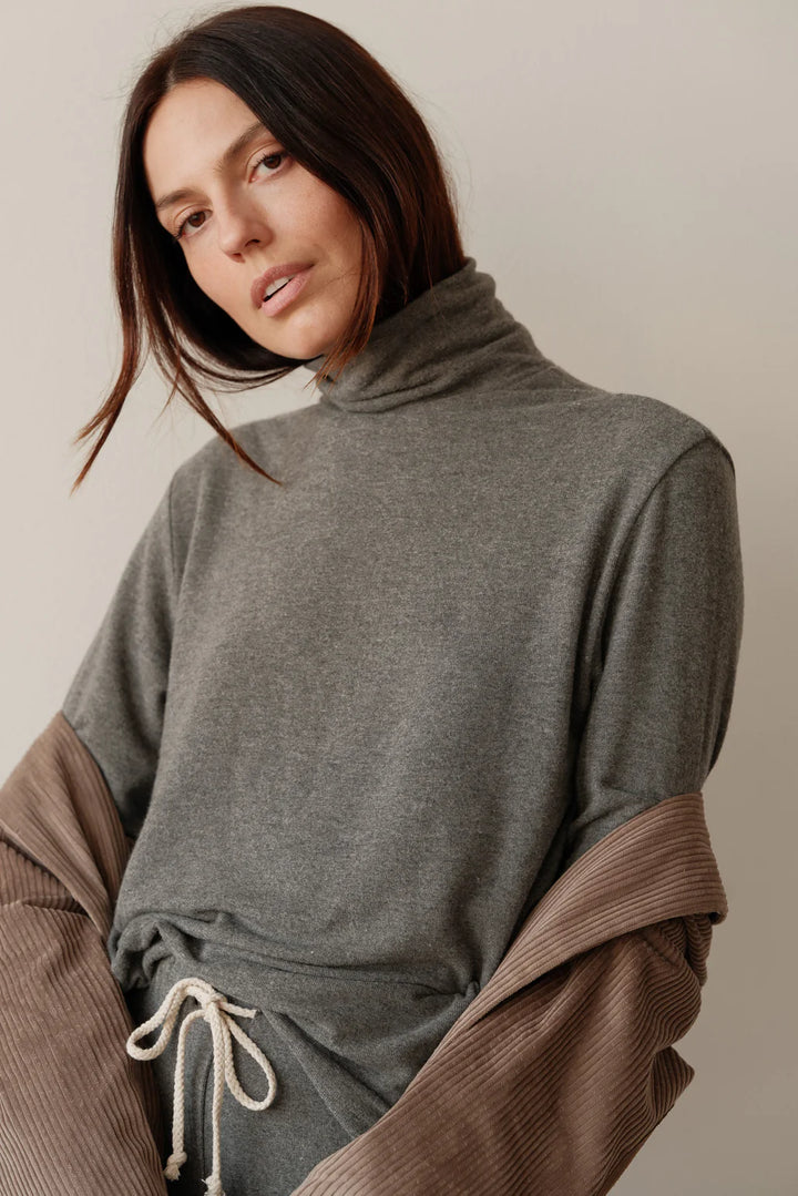 A close up image of a female model posing while wearing the Sweater Turtleneck in Charcoal Grey styled with a corduroy shirt draped over her arms