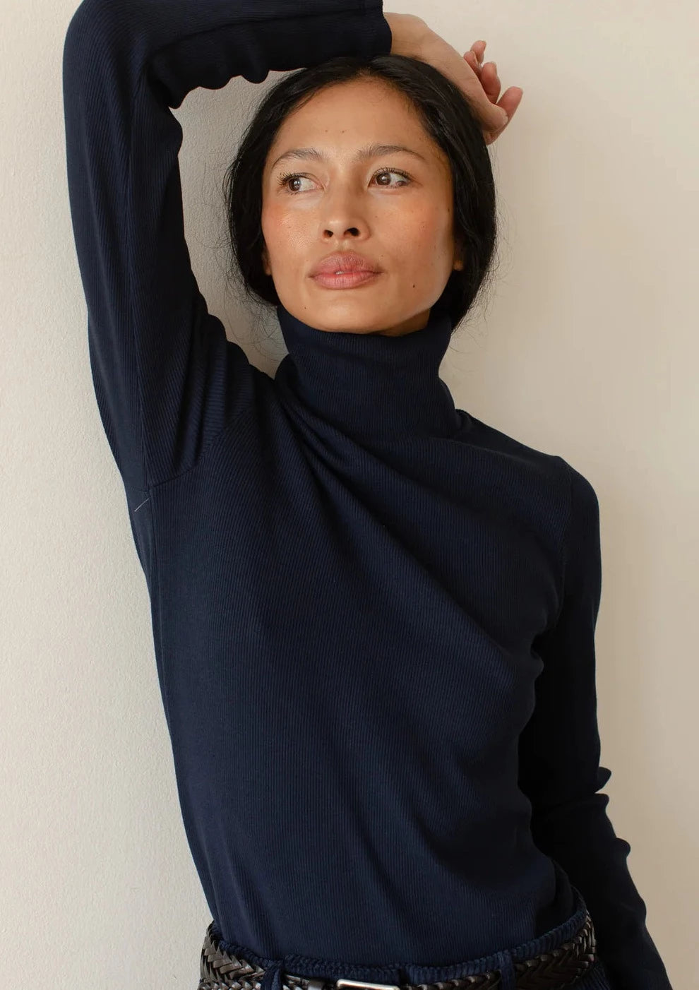 Close image of a model posing with an arm over her head while wearing the Rib Turtleneck in navy