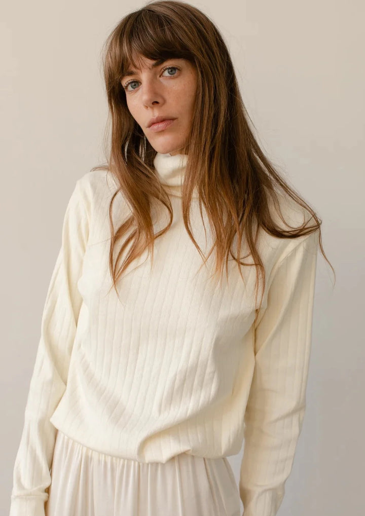 A head shot style image of a female model wearing the Pointelle Turtleneck styled with a matching creme pant 