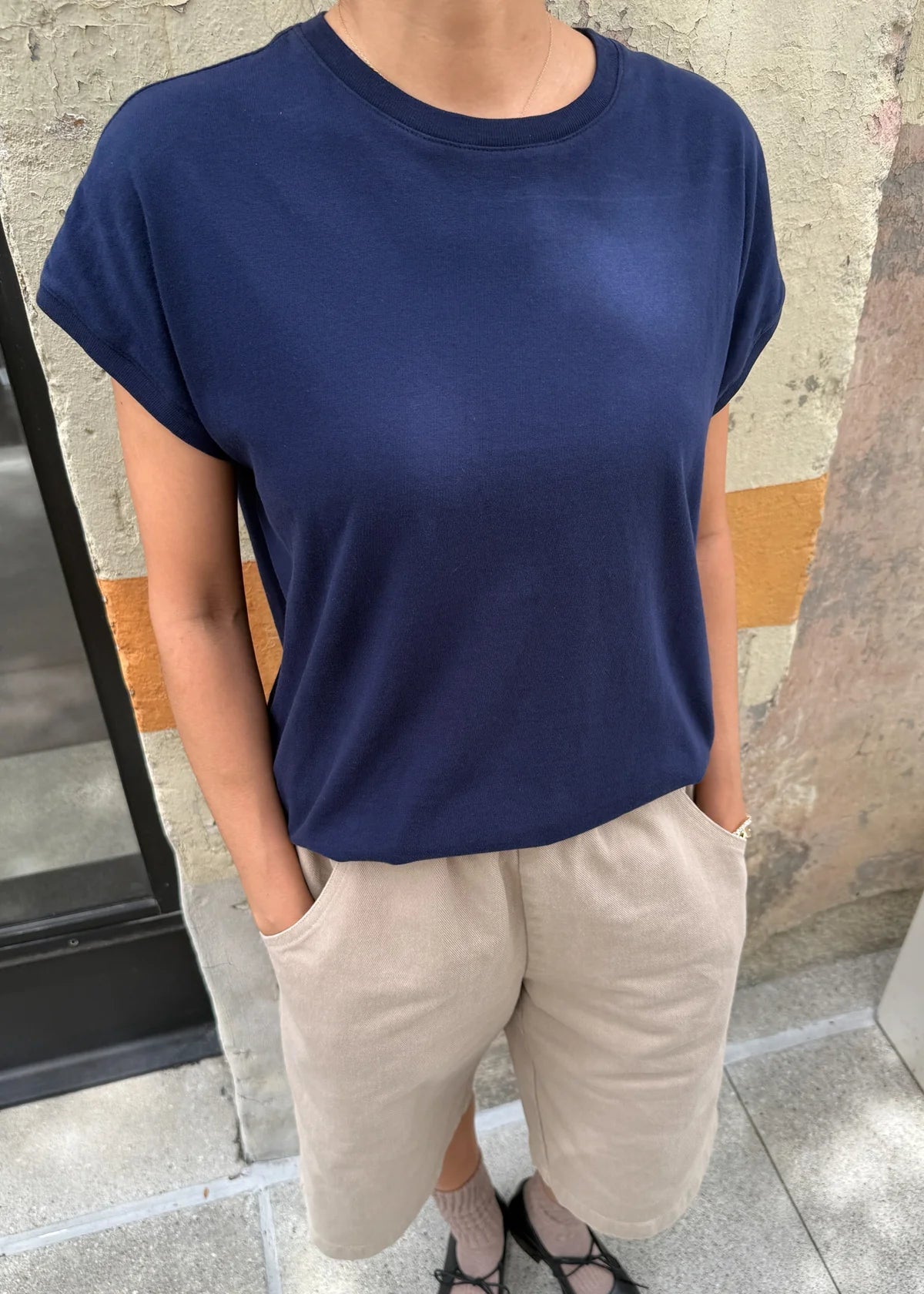 Person wearing a navy blue t-shirt and beige pants standing against a stone wall.