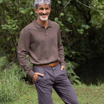 Man wearing a brown sweater and gray pants standing outdoors with greenery in the background