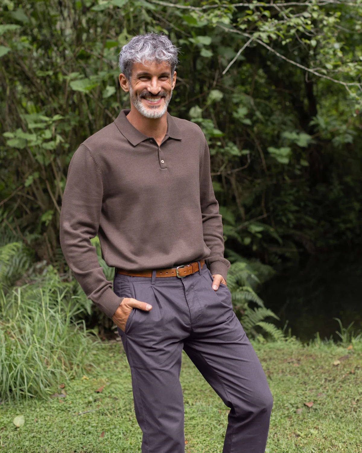 Man wearing a brown sweater and gray pants standing outdoors with greenery in the background