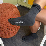 Person wearing black socks with 'LE BON SHOPPE' branding, sitting on a chair with a woven orange ottoman.