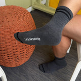 Person wearing black socks with 'LE BON SHOPPE' branding, sitting on a chair with a woven orange ottoman.