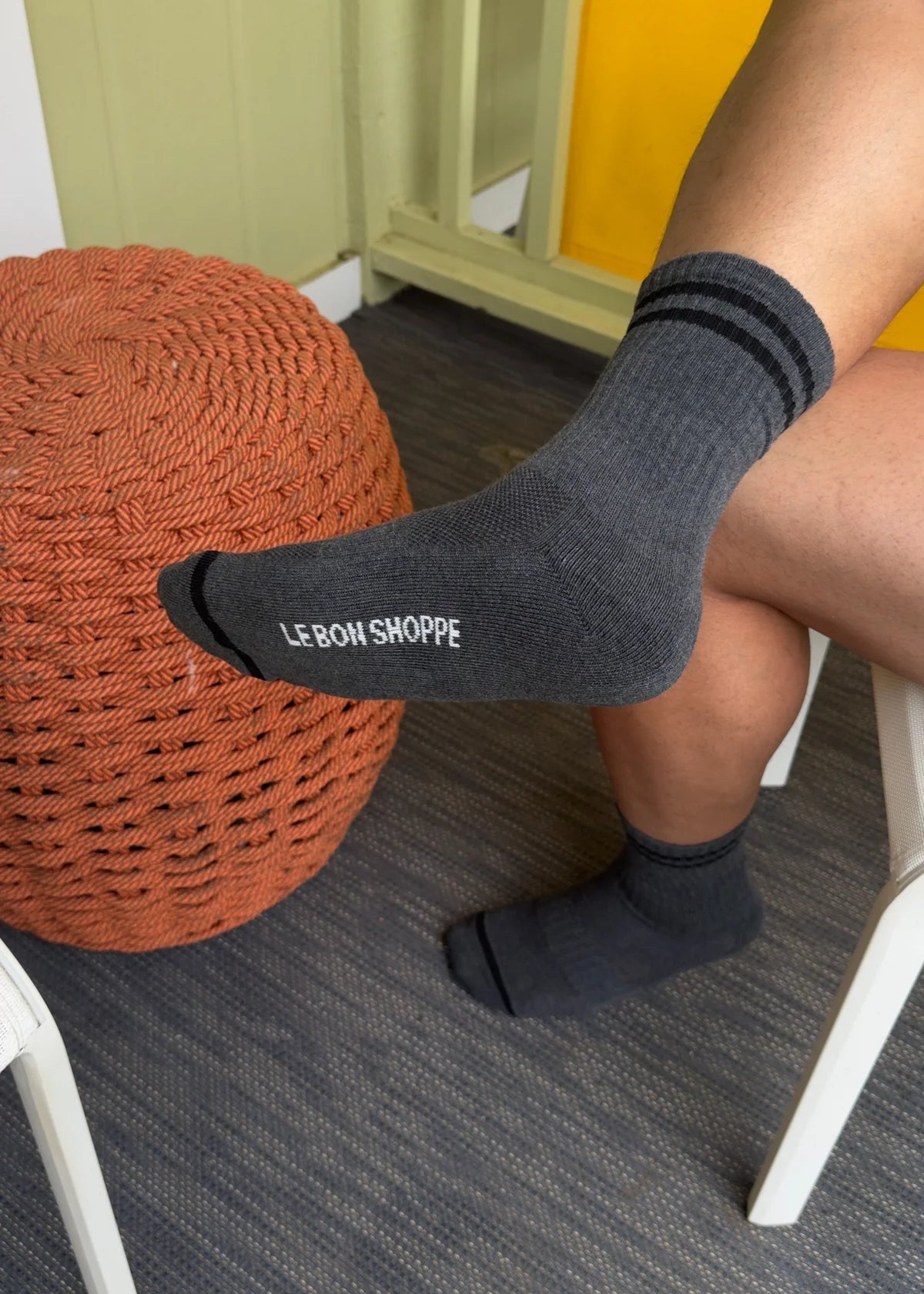 Person wearing black socks with 'LE BON SHOPPE' branding, sitting on a chair with a woven orange ottoman.