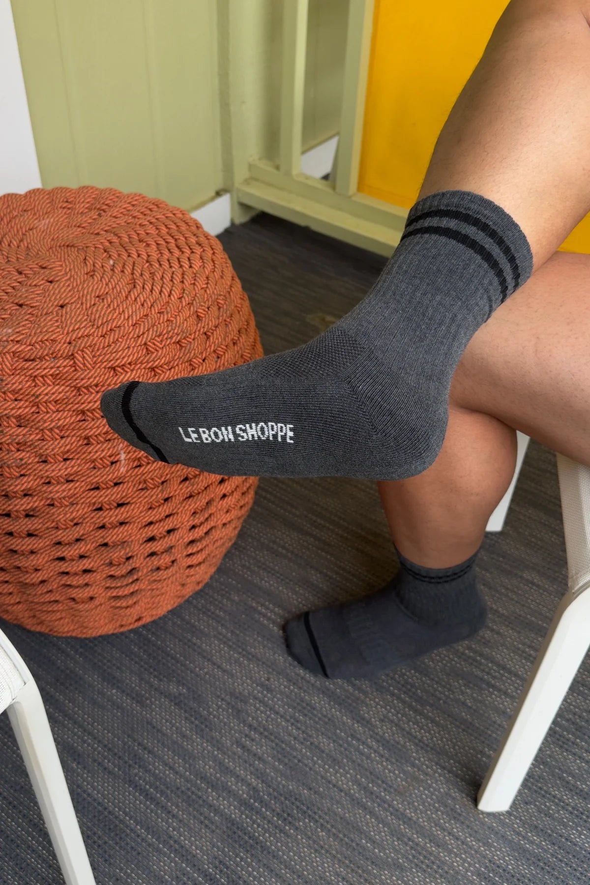 Person wearing black socks with 'LE BON SHOPPE' branding, sitting on a chair with a woven orange ottoman.