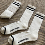 White socks with black stripes and Le Bon Shoppe branding laying on tan carpet.