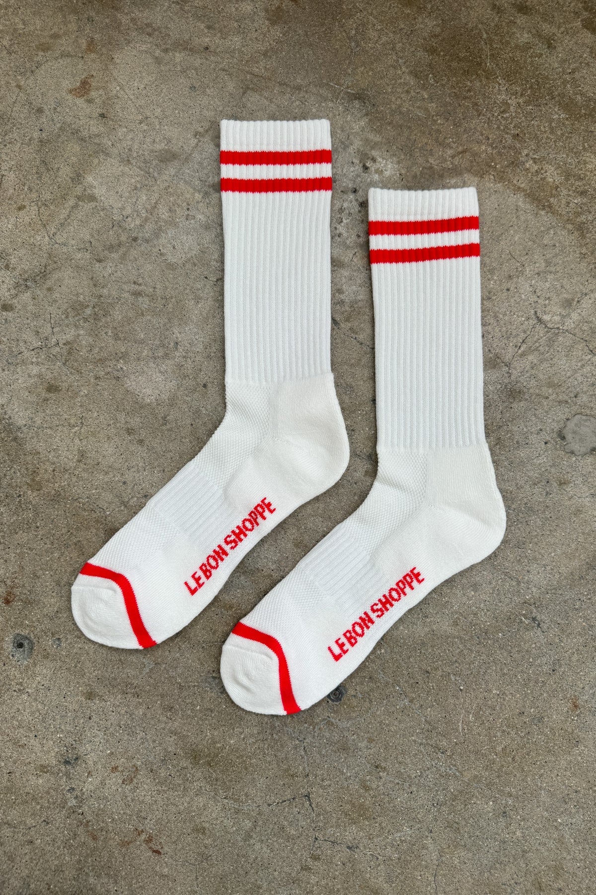 White socks with red stripes and Le Bon Shoppe branding laying on cement floor.