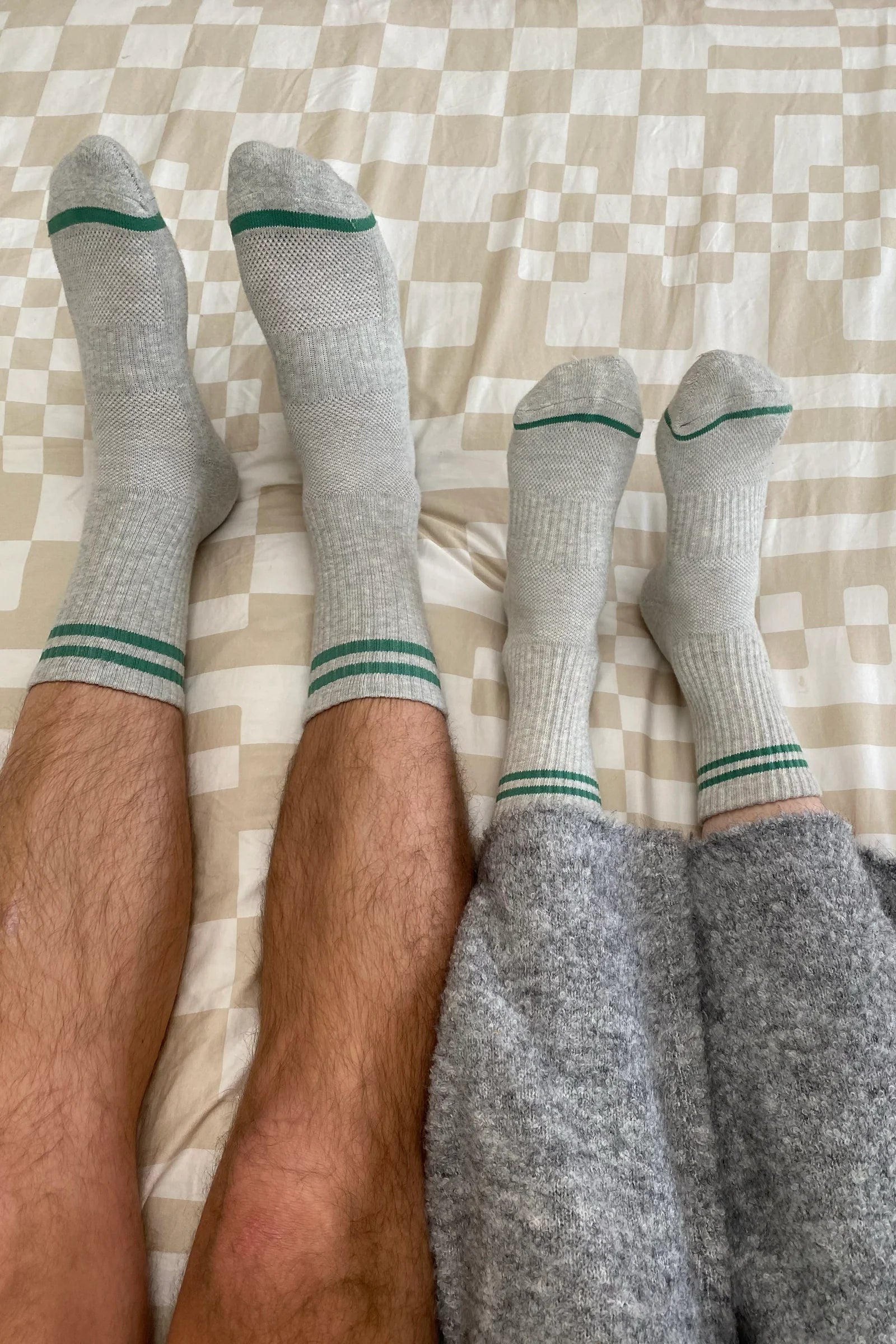 Two pairs of legs wearing gray socks with green accents on a checkered bedspread.