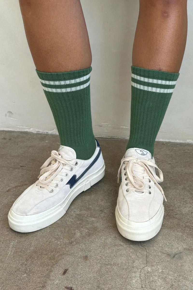Person wearing green socks with white stripes and white tennis shoes on concrete floor