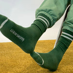 Person wearing green socks with Le Bon Shoppe branding and green sweat pants on yellow carpet