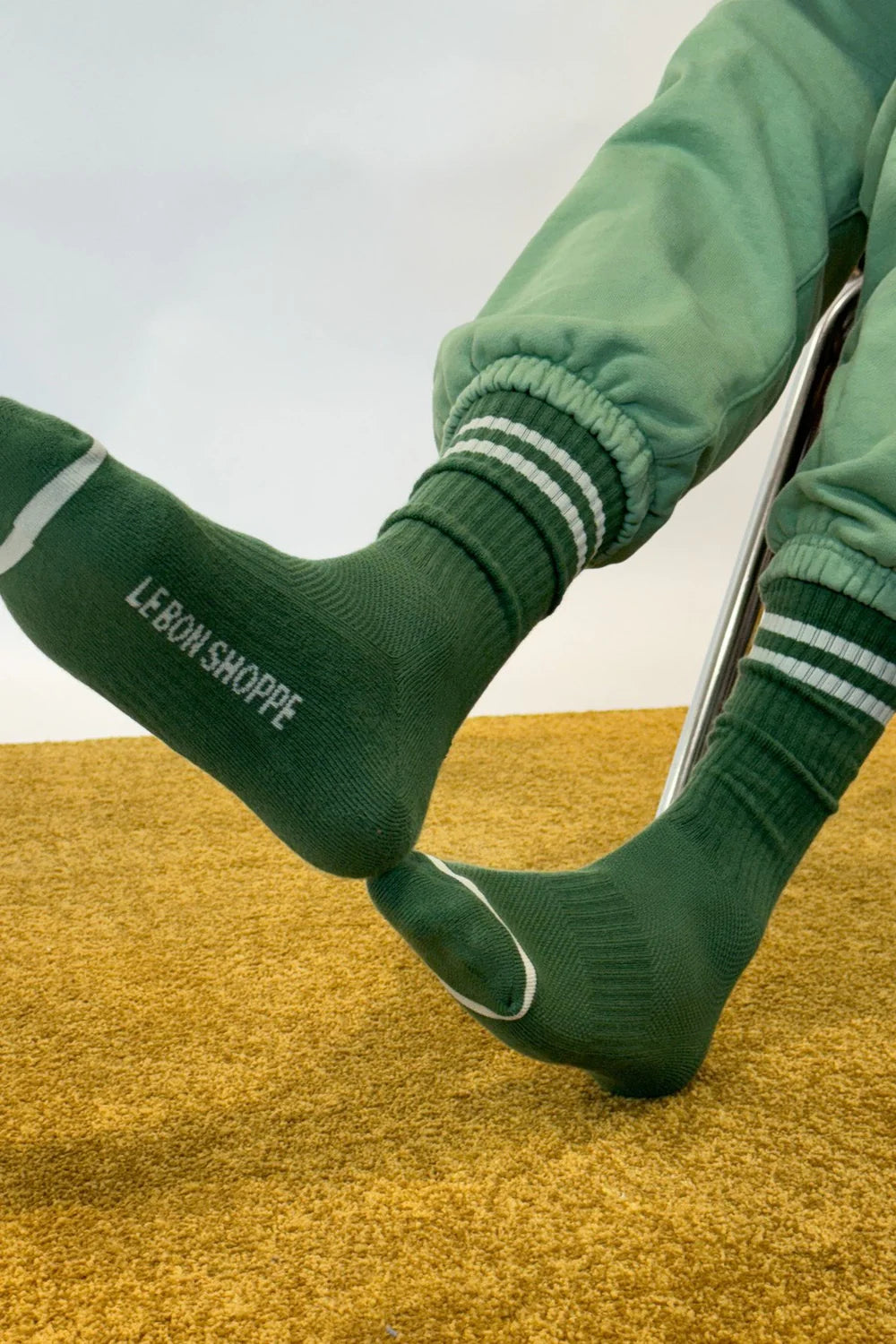 Person wearing green socks with Le Bon Shoppe branding and green sweat pants on yellow carpet