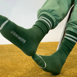 Person wearing green socks with Le Bon Shoppe branding and green sweat pants on yellow carpet