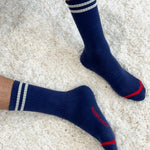 A pair of navy-colored socks with white stripes and red accents on the cuffs, worn on a person sitting on a plush white surface.