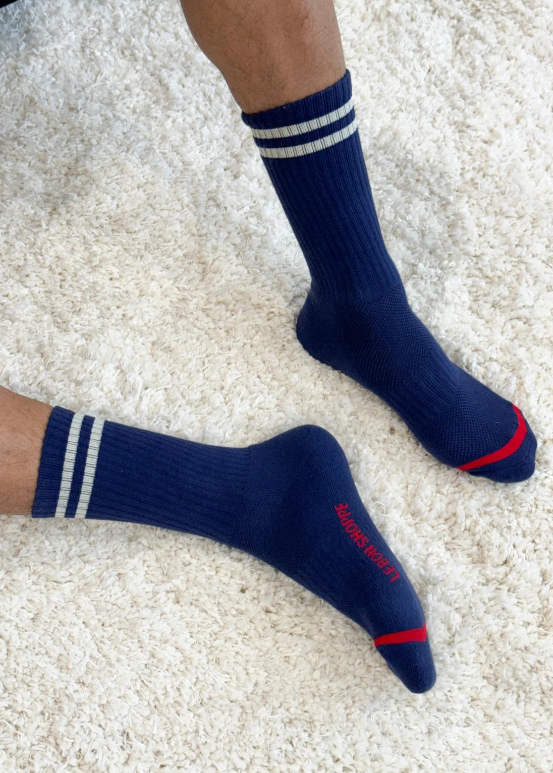 A pair of navy-colored socks with white stripes and red accents on the cuffs, worn on a person sitting on a plush white surface.