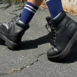 Person wearing navy socks with white stripes and black lace up boots walking on concrete