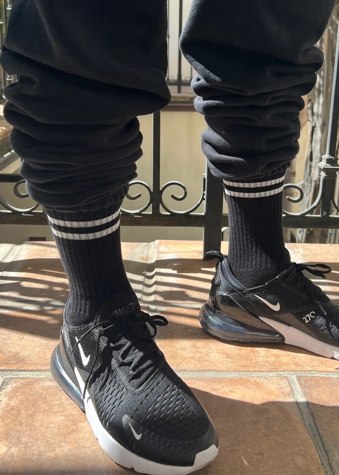 Person wearing black socks with white stripes black sweat pants and black sneakers on a tile floor