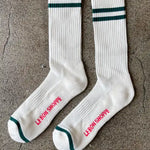 White socks with green stripes and red Le Bon Shoppe branding laying on concrete flooring