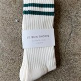 A pair of white, striped, calf-length socks with green and black stripes, displayed on a concrete surface with a tag showing 'le bon shoppe'.