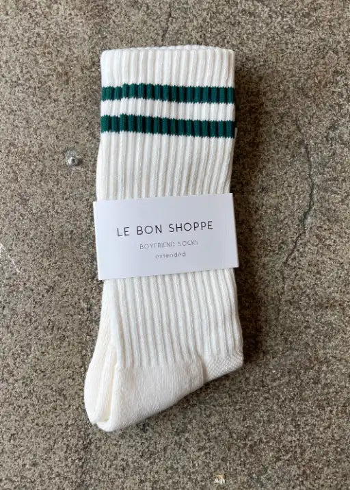 A pair of white, striped, calf-length socks with green and black stripes, displayed on a concrete surface with a tag showing 'le bon shoppe'.