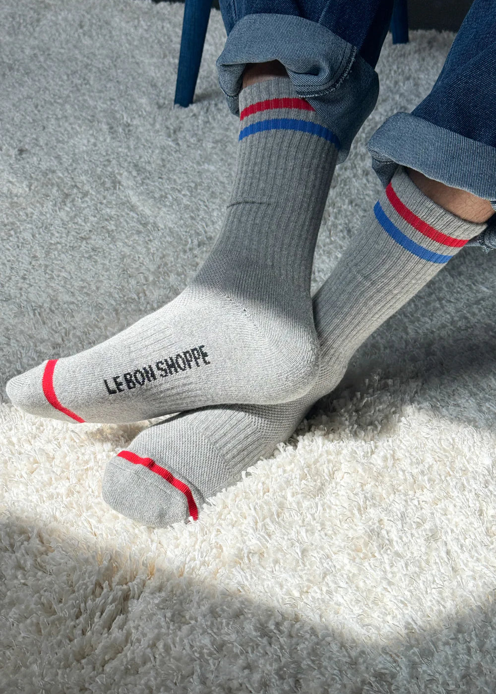 Person wearing grey socks with red and blue stripes and Le Bon Shoppe branding and jeans on shaggy carpet in a spot of sun