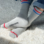 Person wearing grey socks with red and blue stripes and Le Bon Shoppe branding and jeans on shaggy carpet in a spot of sun