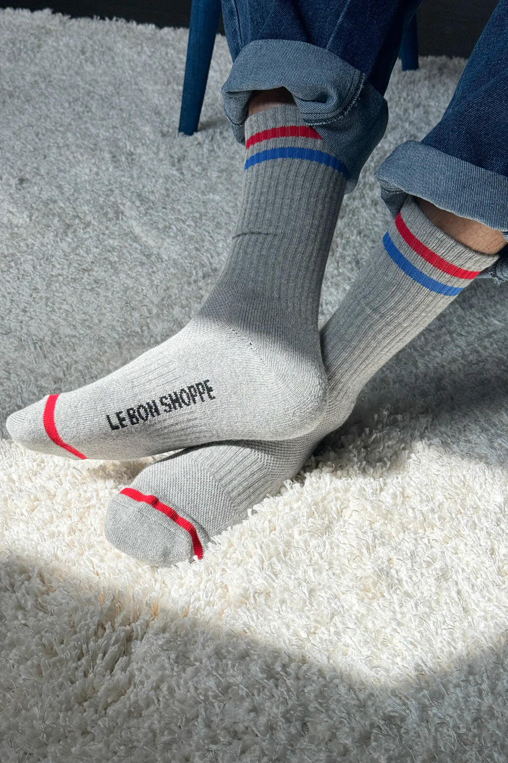 Person wearing grey socks with red and blue stripes and Le Bon Shoppe branding and jeans on shaggy carpet in a spot of sun