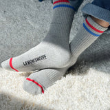 Person wearing grey socks with red and blue stripes and Le Bon Shoppe branding and jeans on shaggy carpet in a spot of sun