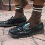 Black leather loafers worn with brown socks on a brick pavement.