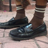Black leather loafers worn with brown socks on a brick pavement.