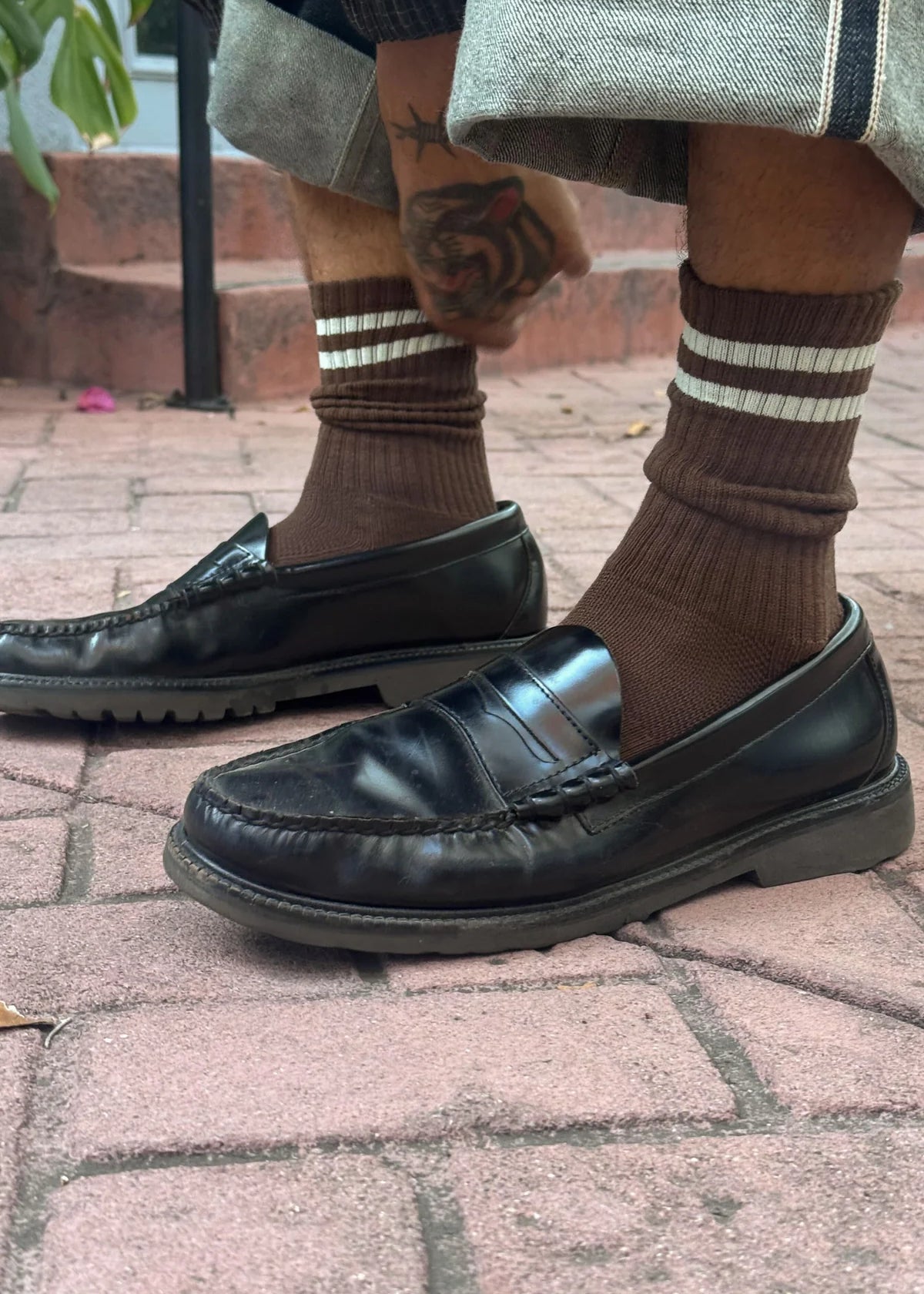 Black leather loafers worn with brown socks on a brick pavement.
