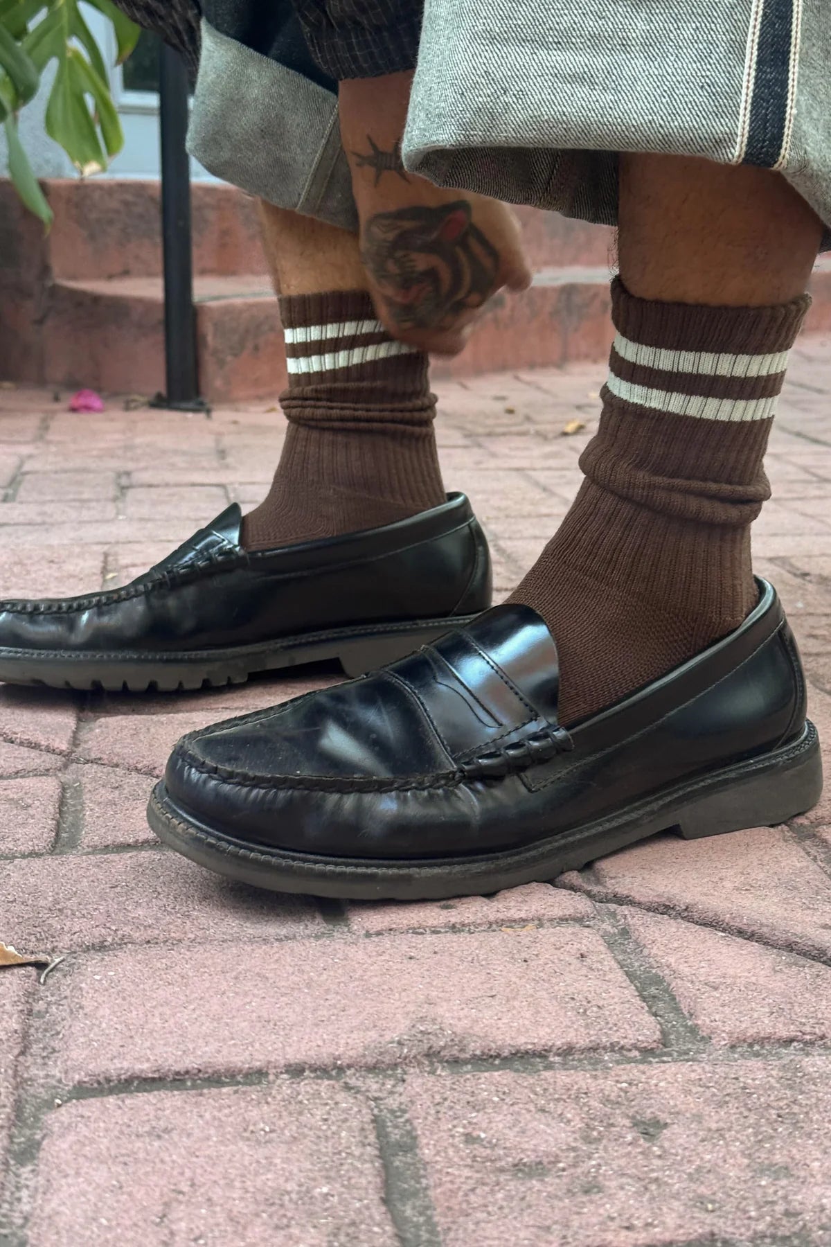 Black leather loafers worn with brown socks on a brick pavement.