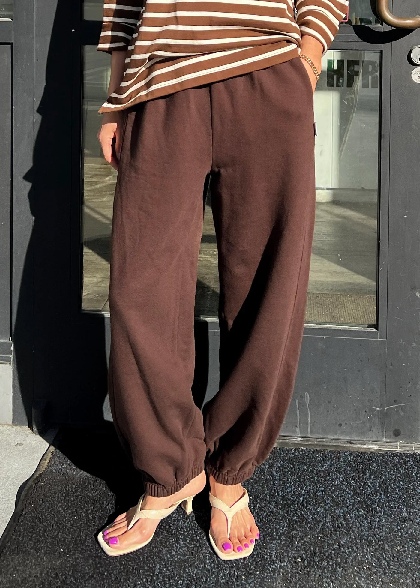 Person wearing brown striped shirt and brown pants standing in front of a glass door.
