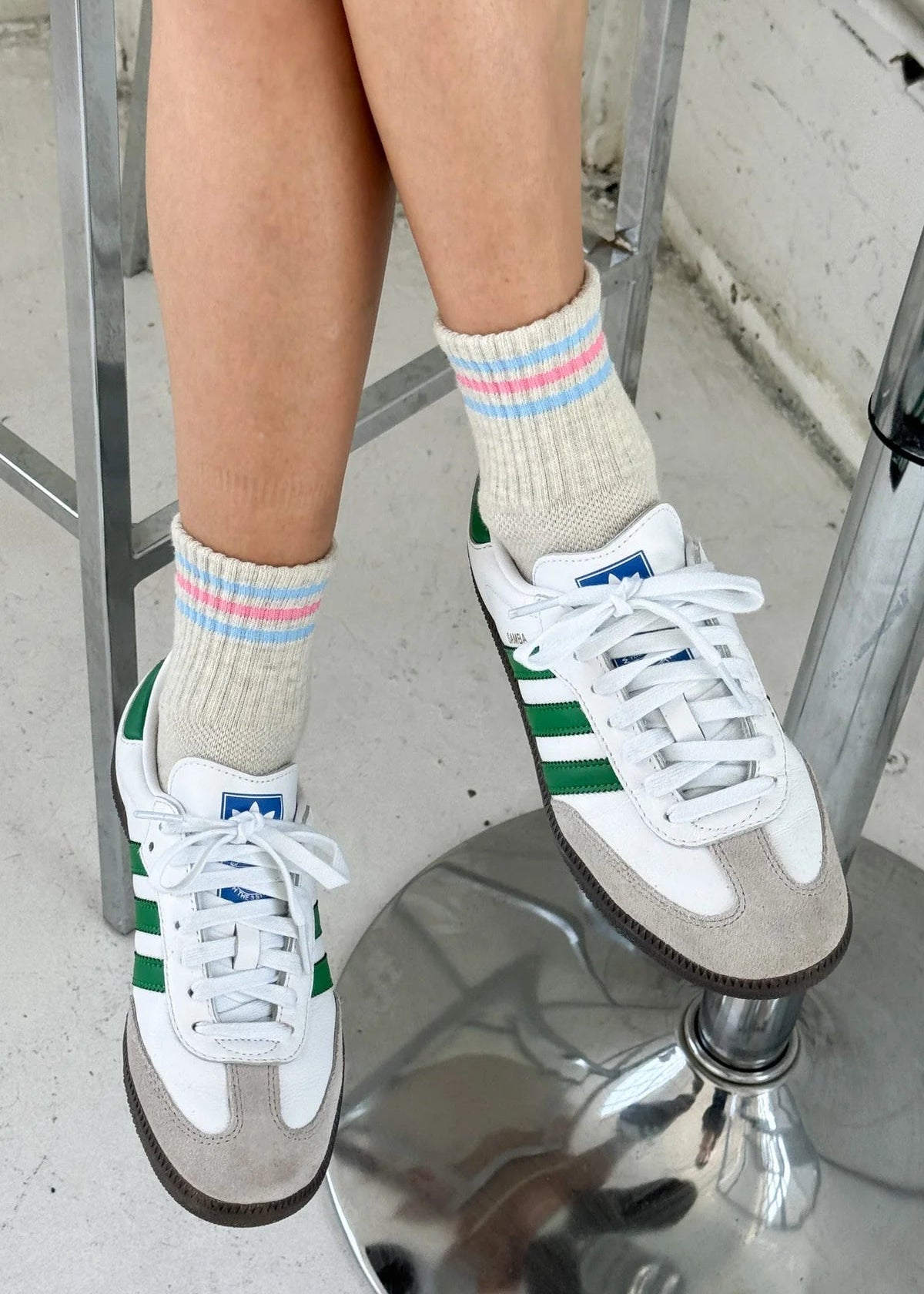 Person wearing white sneakers with green accents and colorful striped socks on a metal stool.