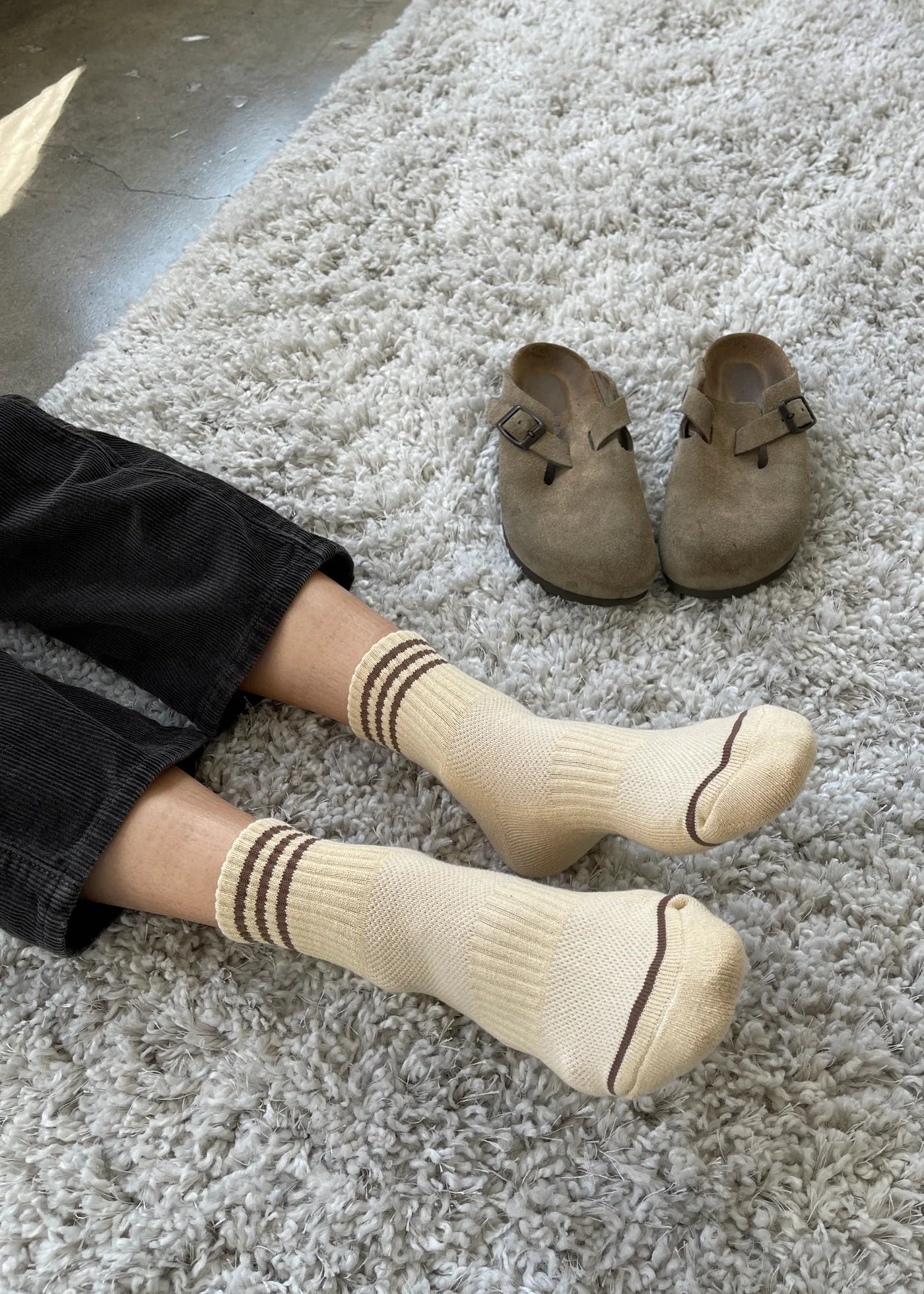A pair of yellow ankle crew socks with triple contrast stripes, displayed on a person's feet protruding from a light-colored blanket, alongside a pair of brown sandals.
