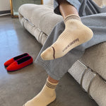 A person in grey pants and yellow socks sitting on a taupe couch with red shoes next to them on the floor.