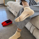 A person in grey pants and yellow socks sitting on a taupe couch with red shoes next to them on the floor.