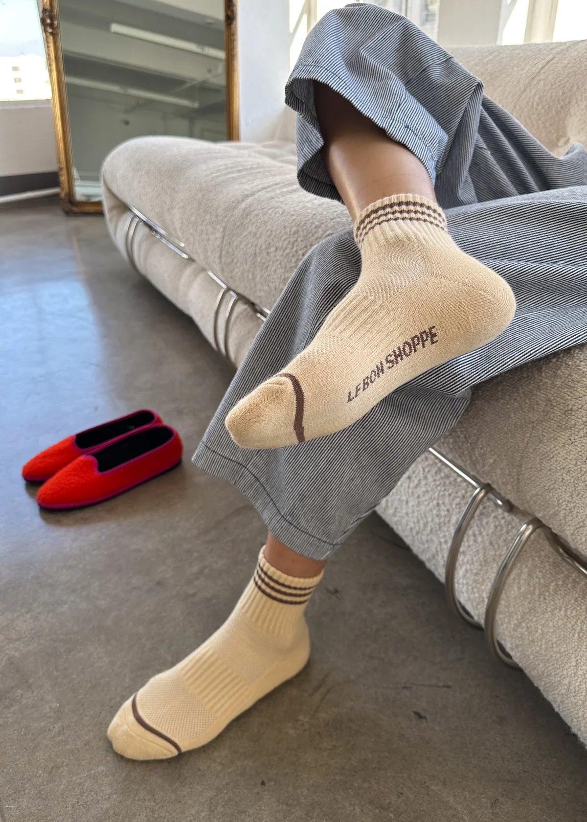A person in grey pants and yellow socks sitting on a taupe couch with red shoes next to them on the floor.