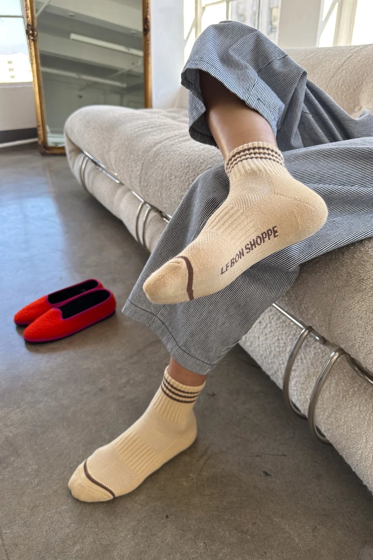 A person in grey pants and yellow socks sitting on a taupe couch with red shoes next to them on the floor.